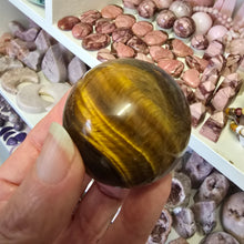 Load image into Gallery viewer, Hand holding a tiger&#39;s eye crystal ball with a display of various crystals in the background.
