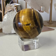 Load image into Gallery viewer, Tiger&#39;s eye crystal ball on a stand with crystals in the background
