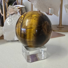 Load image into Gallery viewer, Tiger&#39;s eye crystal ball on a stand with crystals in the background
